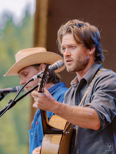 Close-up of Jamestown Revival musicians performing live with microphones and acoustic guitars, one wearing a cowboy hat, captured by Joseph Mayden during an outdoor concert.