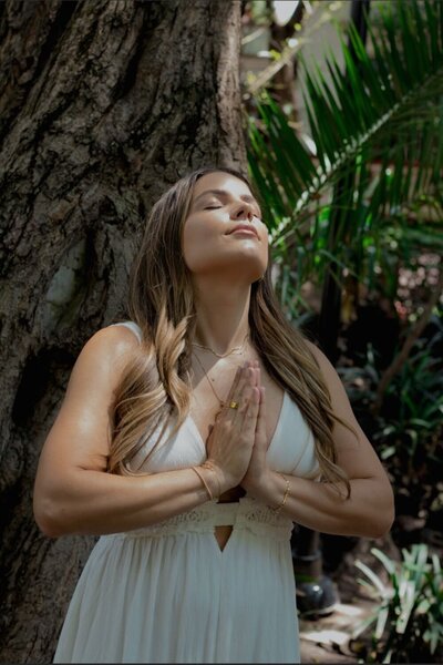 Natalie Bernacchi standing in prayer pose under a tree, eyes closed, connecting to nature and grounding her energy for Reiki work