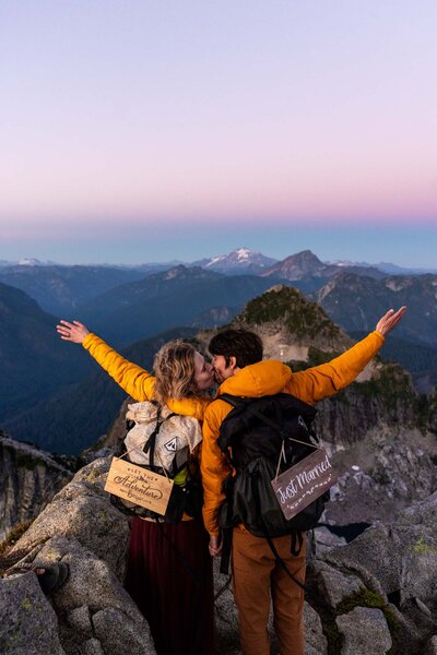 A couple kissing with their arms in the air at sunset. They are standing on top of a mountain wearing backpacks with elopement signs.