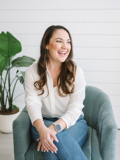 Woman sitting in a teal chair with a plant behind her and smiling during her brand photoshoot