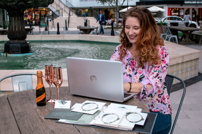 woman sitting at a table outdoors working on a laptop with wedding invitations scattered on table