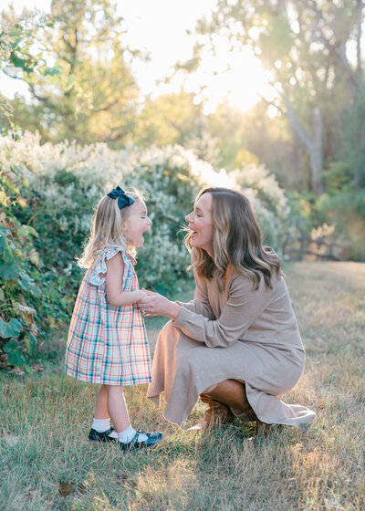 Mom crouched down holding the hands of her toddler daugher smiling at each other surrounded by green foliage and a golden sunset- Maegan R Photography