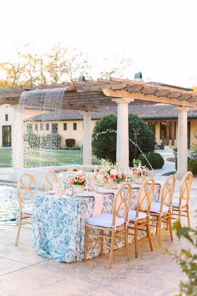outdoors reception table with floral blue tablecloth and peach and white bouquets on top, with natural wood and white chairs and a water fountain in background.