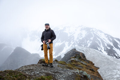 Elopement photographer stands on a rugged mountain ridge, holding a camera with misty peaks and a glacier in the background.