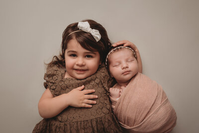 20 month big sister in brown dress laying on back holding newborn baby sister wrapped in pink fabric swaddle
