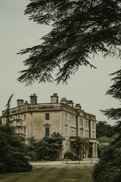 A stately country manor stands tall amidst lush green trees, its historic architecture exuding timeless elegance in the English countryside.