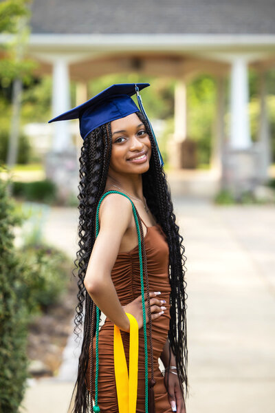 African American girl wearing a brown fitted short dress, graduation cap posing with awards and cords