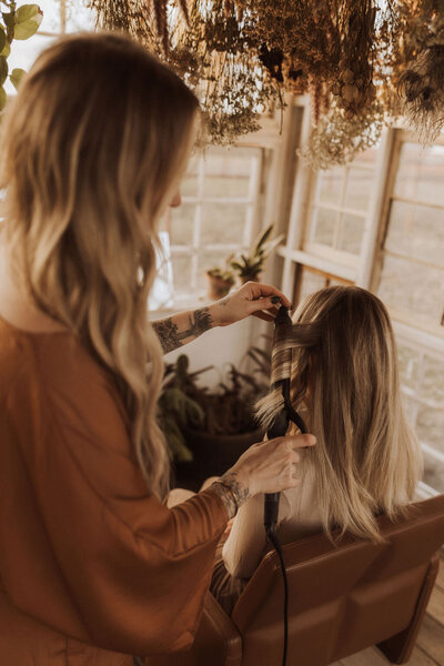The color and hair extensions professional is working on the hair of a blonde client sitting in a brown chair. The salon is decorated with green plants and flowers on the walls