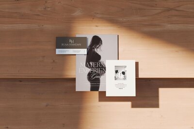 Flat lay of Ruaa Hamdan Photography branding set including maternity guide booklet and promotional cards on wooden surface in sunlight