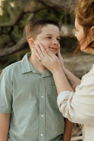A woman gently holds a young boy’s face in her hands while they stand outdoors, both looking at each other.