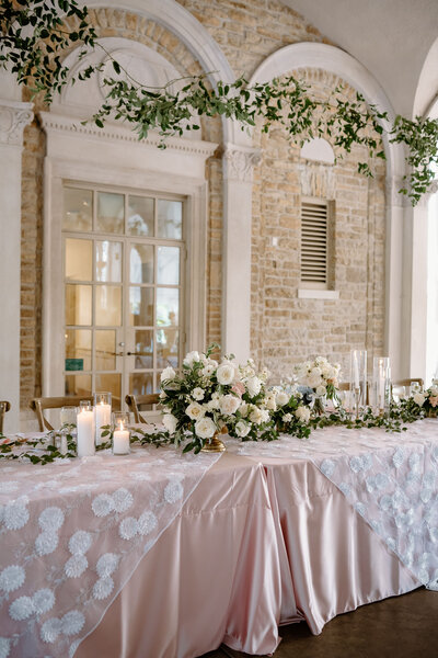 Romantic sweetheart table with blush linens, white florals, candles, and greenery.