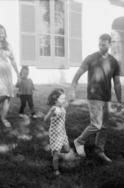 Candid black and white film image by a family photographer in Richmond, VA of a family walking through the yard together. The little girl runs ahead while her parents and younger sibling follow. Natural movement and timeless tones.