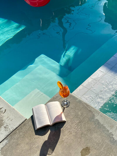 A glass of cocktail next to an open book by a pool