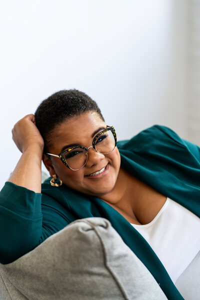Career change consultation expert wearing a green branded blazer and reading glasses, sitting on a couch with a kind gesture as her arm gently touches her head.