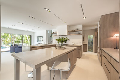 Bright and cream colored kitchen showcasing Sarah's interior design. The focus is a large cream coloured island bar, with a low-back bar stool tucked in. On the island is a vase with green foliage. The kitchen has light wood open shelves.