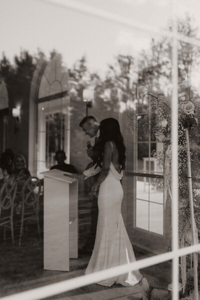 Black and white photo taken from outside a window, showing bride and groom at the podium with groom holding a microphone.