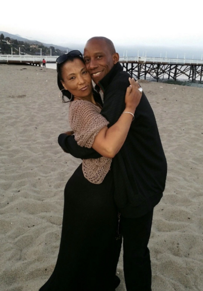 Image: Regina and Stan embracing cheek to cheek. Standing on the beach. Regina is wearing black summer dress and beige knit crop sweater. Stan is wearing a casual black jacket and pants. Both smiling. The Malibu pier is in the background.
