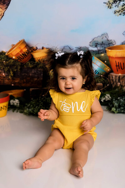 Baby girl in yellow bee themed romper smiles during adorable first birthday portrait with honey pot decorations