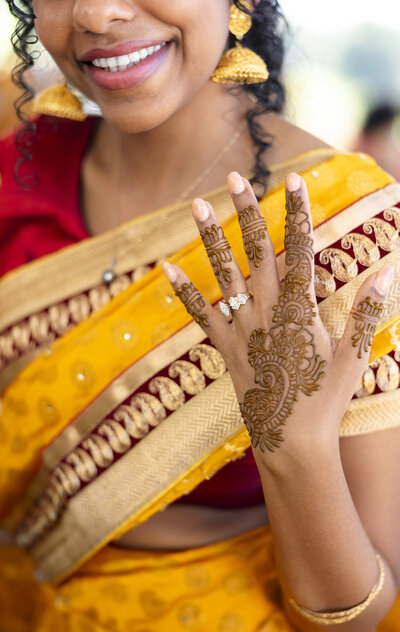 A woman in a vibrant yellow and red saree smiles, showing intricate henna designs on her hand. She wears gold earrings and a diamond ring on her finger. The background is softly blurred, highlighting the detailed patterns.