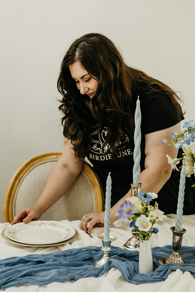 A woman wearing a shirt that says Birdie June sets a plate on a table with candles and flowers, representing wedding management services. 