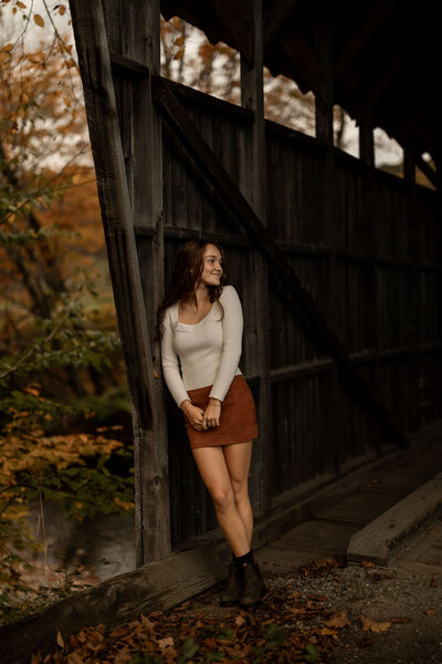 A woman in a white top and brown skirt leans against a wooden structure, smiling and glancing aside. The scene, captured by a documentary photographer in New England, features autumn leaves adorning the path and trees cloaked in orange and yellow, exuding a warm, rustic charm.