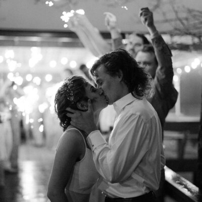 photo of couple embracing a kissing. The woman is on the left, wearing a flower crown and white dress. The man is on the right, holding her face, wearing a white collared dress shirt.