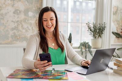 Photo of woman sitting at a desk multitasking with a calendar, computer and phone, woman is smiling at phone.