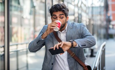 Stressed businessman drinking coffee and checking watch while holding a phone, rushing through a city street.