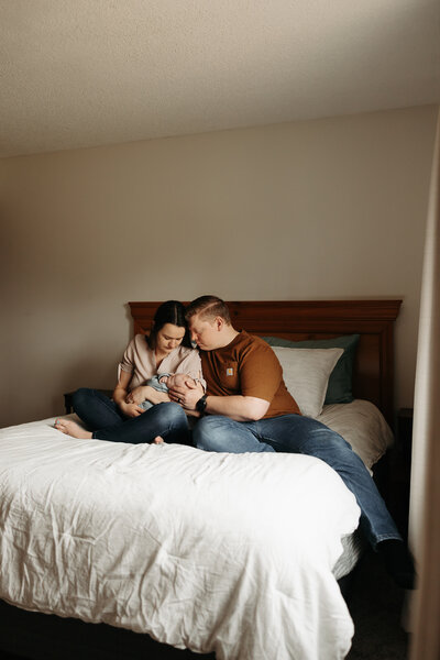 Woman and man sitting on bed holding newborn baby and looking at him