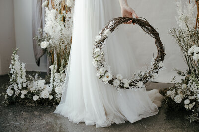 Detail of a  bride in a tulle gown holding a wedding wreath of dried white flowers .