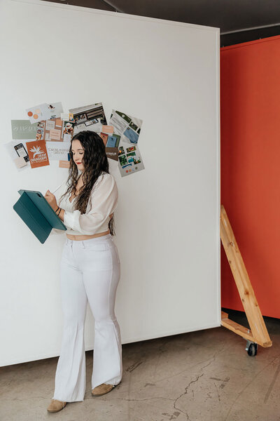 Taysha working on her tablet leaning agains a white wall with a moodboard hanging behind her.
