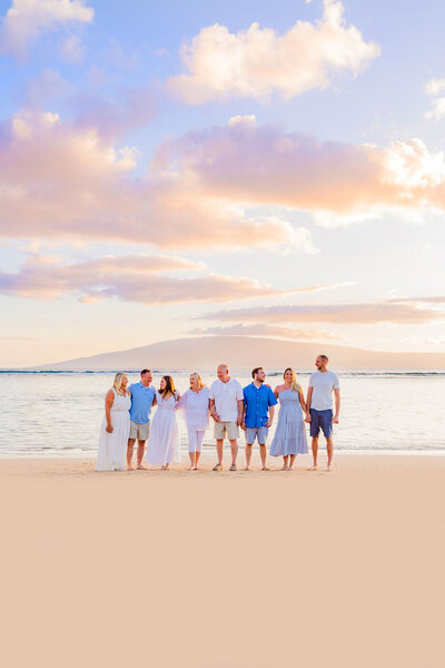 Woman in white off-shoulder dress holds her children's hands on either side as they dip their toes into the water at the beach in West Maui