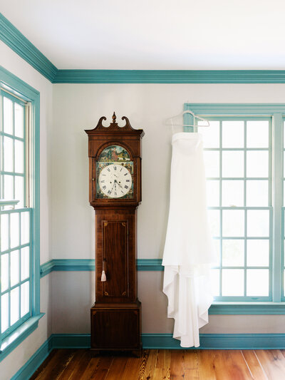 A simple, down-to-earth wedding dress hangs gracefully next to a vintage grandfather clock at the Merrimon-Wynne House, captured by Jessica Leigh Photography.