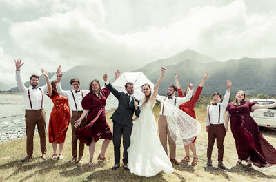 A bride and groom stand outside with guests, all smiling and raising their arms in celebration during their wedding photoshoot.