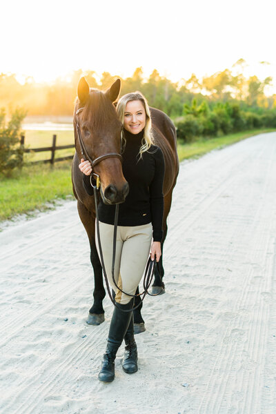 girl standing on a dirt road holding a horse with the sun setting behind them