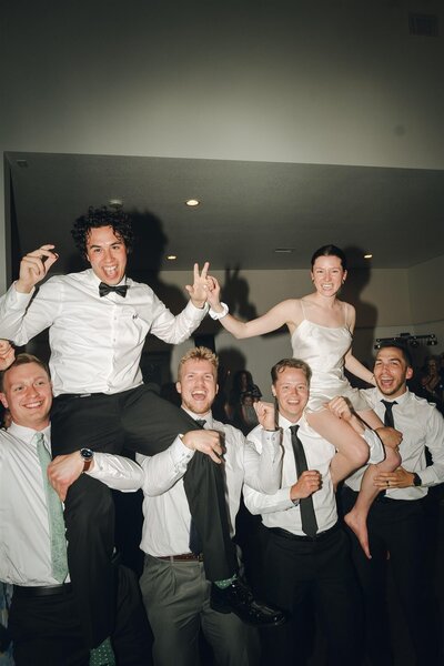 Groomsmen hold bride and groom up on their shoulders during raucous wedding reception.