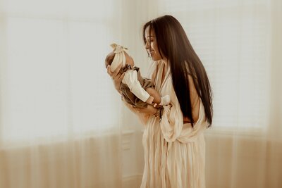 In a sunlit room, captured by a Macon newborn photographer, a woman with long hair gazes lovingly at her baby, both smiling. The little one is dressed adorably in a headband and onesie.
