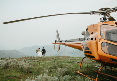 A couple dancing on top of a mountain in their wedding clothes, the bride is wearing a white dress and the groom is wearing a black suit, next to an orange helicopter