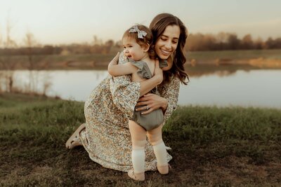 A woman kneels on grass near a body of water, hugging a young child who is standing and wearing a bow and romper—captured beautifully by a Macon GA photographer.