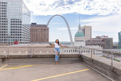 Photograph of a woman leaning against a fence with the St. Louis Arch, Hyatt, and Courthouse in the background.