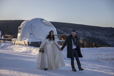 Bride and groom walk hand in hand through the snow at an Alaska elopement, with a modern glass-domed igloo behind them. The bride wears a flowing wedding dress with a warm cape, while the groom is dressed in a dark winter coat, capturing the magic of a winter elopement under the northern sky.