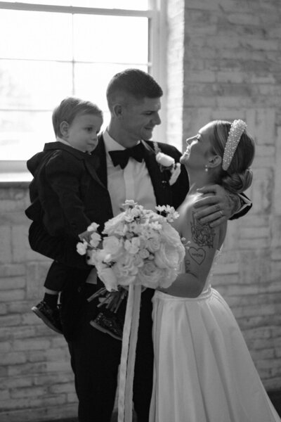 A bride and groom smiling at each other while holding their baby boy.