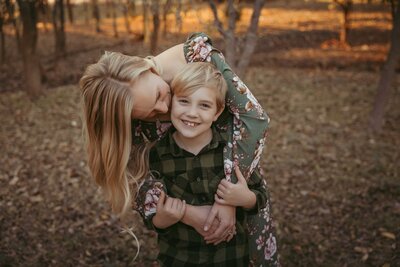 A mother's love captured in a heartfelt moment as she leans forward and tenderly kisses her son on the cheek. They are both standing in the middle of a wooded area with crunchy brown leaves on the ground.