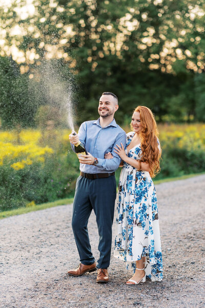 Couple walks along path at The Willowwood Arboretum popping a bottle of champagne.
