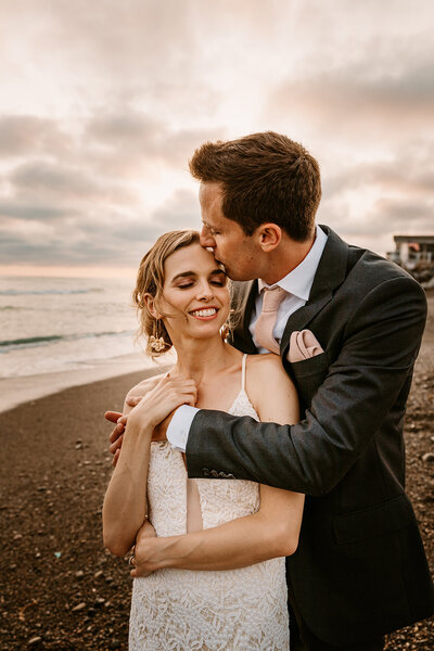 groom kisses bride on forehead on beach