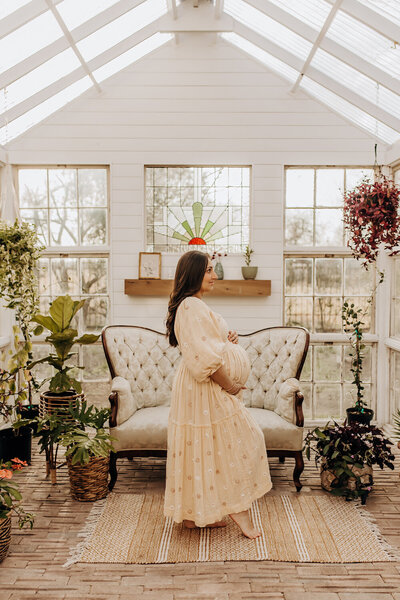 A San Antonio greenhouse mini session for a mother to be who is holding her bump in a flowy, cream dress.