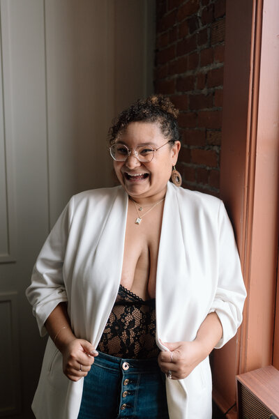 Ashley smiling away from the camera,  in a white blazer and  black lace body suite.