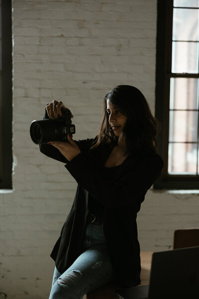 A New England Wedding Photographer in a dark jacket stands near a window, holding a camera and peering through the viewfinder. The room's white brick walls embrace the streaming natural light as she remains focused and intent on capturing the perfect shot.