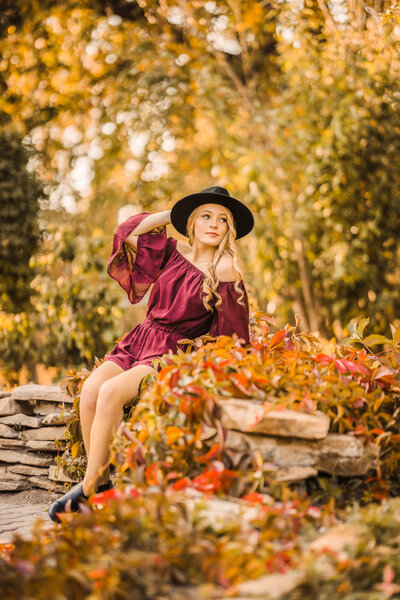 senior girl in a dark red dress and black hat sitting in a flower garden during the fall season