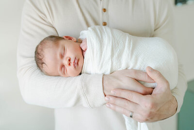 baby in white onsie is held in Father and siblings arms during a newborn photo session with AnneMarie Hamant